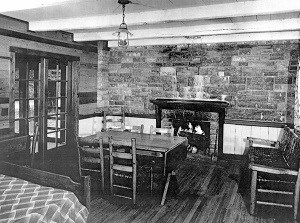 black & white photo of interior of cabin with stone fireplace wall at back and table and chairs at center