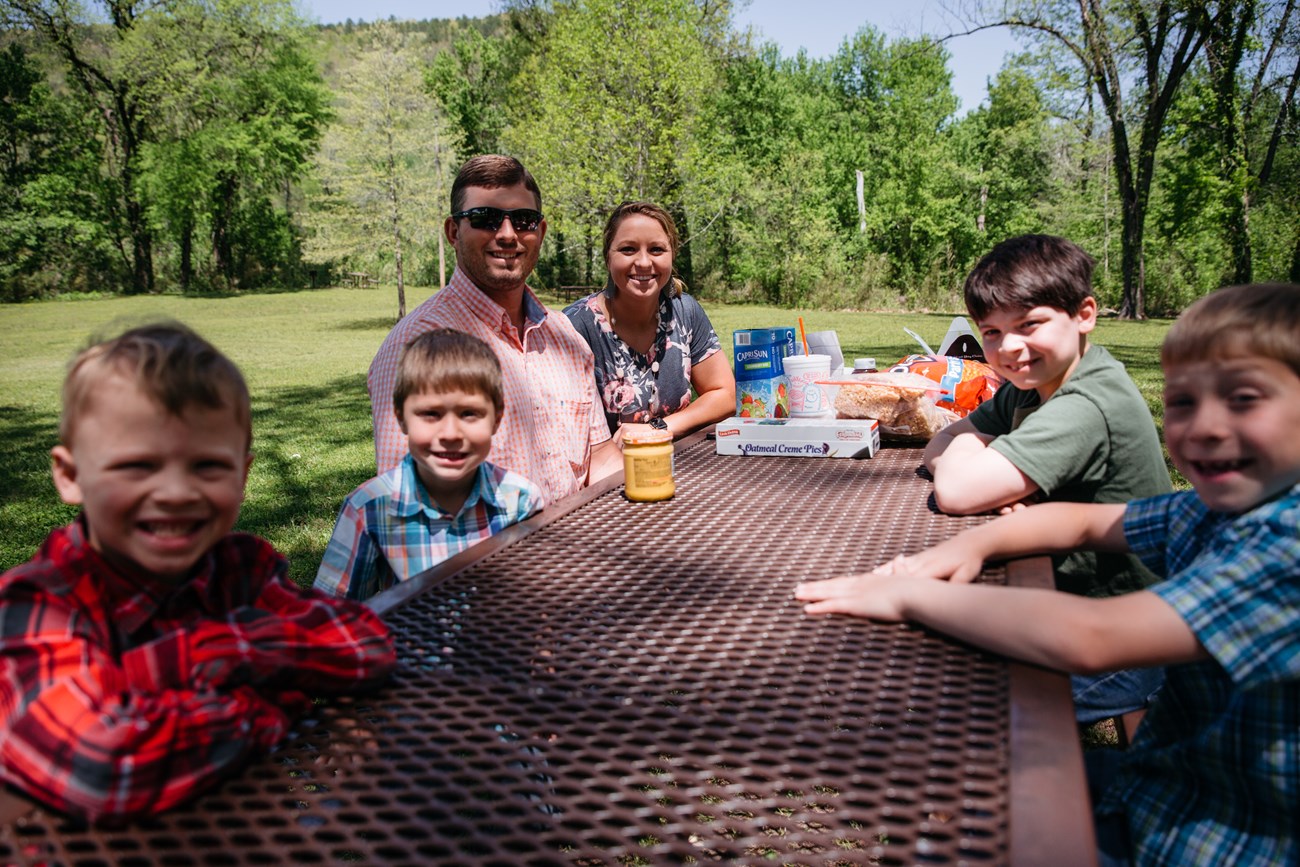 A mother, father, and four boys sitting at a picnic table with snacks.