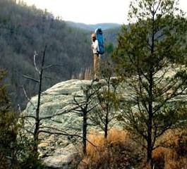 Man wearing large backpack standing on rocky cliff with view of forested valley.