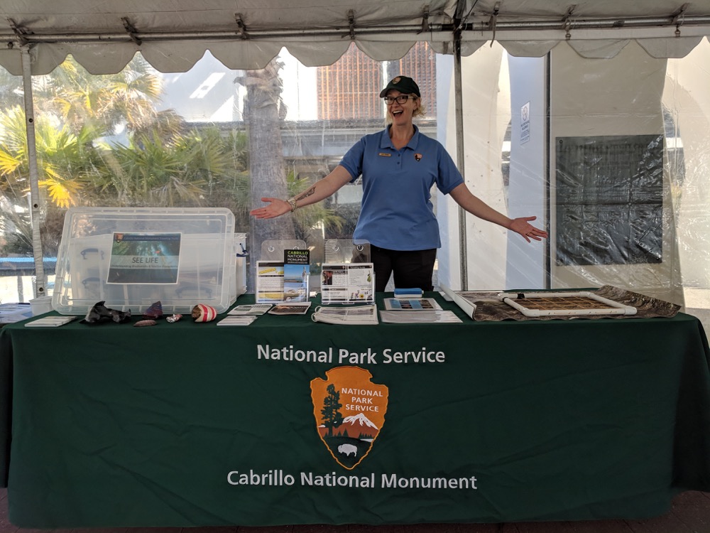 Scientist Samantha Wynns stands behind her table of 3D-printed tidepool creatures and a tidepool monitoring activity, ready to engage students.