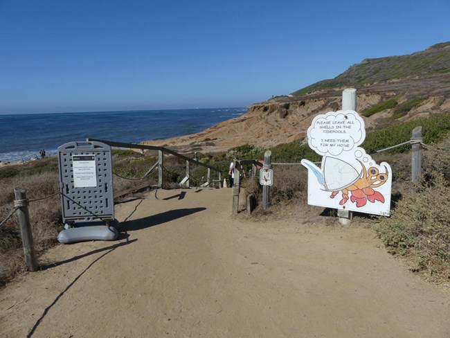 A sign with a drawing of a crab along a dirt path leading to stairs. The ocean is in the distance.