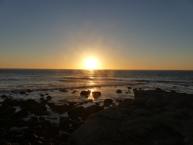The sun sets into the ocean with a rocky beach in the foreground.