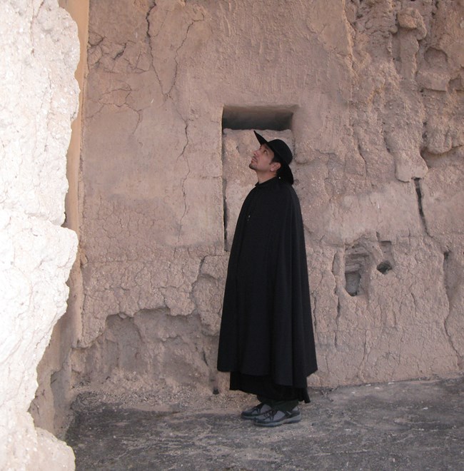 Park Superintendent dressed in dark historical robes  and a dark brimmed hat stands inside the Great House ruins as part of the film shoot