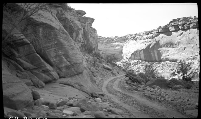 Black and white photo of narrow dirt road winding through narrow, rocky canyon.