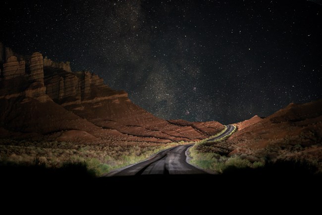 Blacktop road at night, curving between red hills and cliffs, underneath the Milky Way.