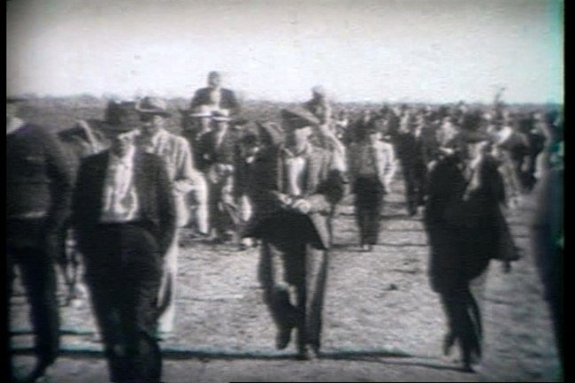 Black and white historic image of gentlemen dressed in suits and hats walking on a dirt road to the Magnolia Horse races.