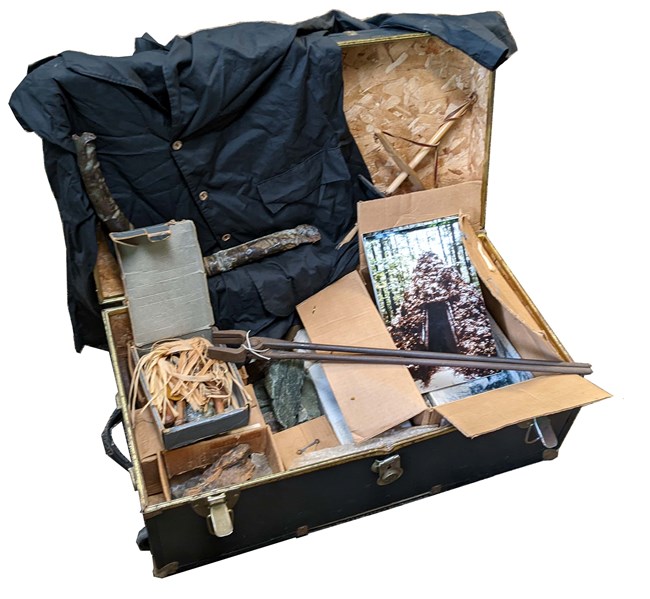 Black trunk holding historic artifacts including a shirt, photo of a hut, and iron tongs