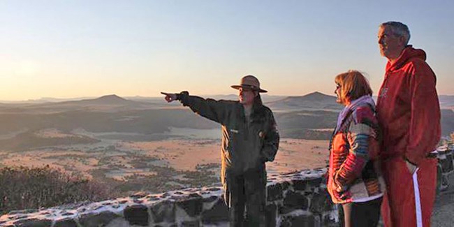 Ranger and two visitors stand at an overlook in early morning light.