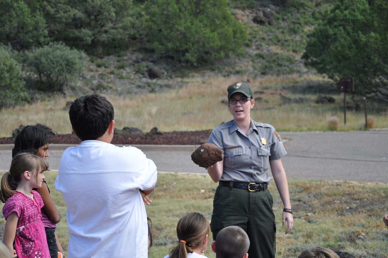 A park ranger is giving a presentation.  She stands facing the camera while holding a rock outward toward a group of five children who are focused on her explanation.  The ranger is wearing a classic ballcap, button-up shirt with name tag and badge.