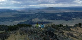 Color photograph from top of Capulin Volcano of mountains and mesas in distance and hikers on trail in foreground.