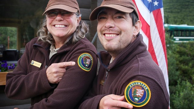Two smiling people point the National Park Service volunteer patch on the left shoulder of their jacket.