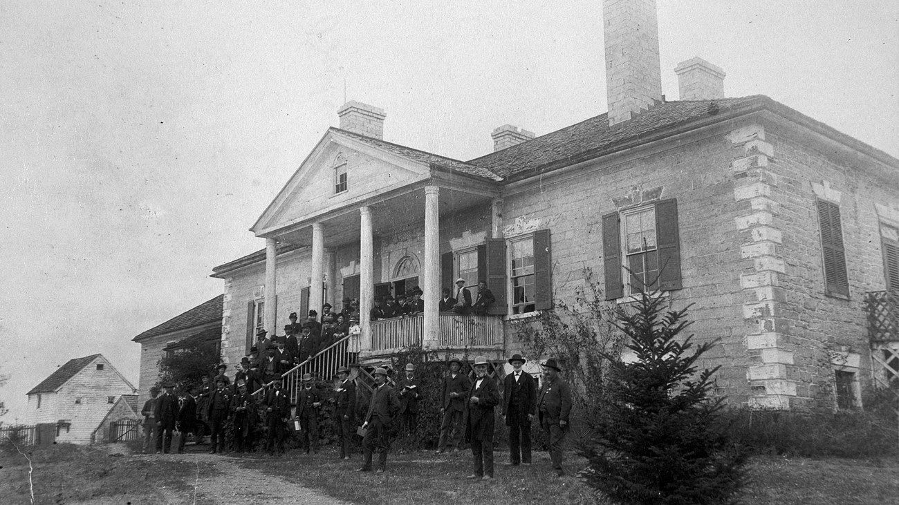 An 1883 photo captures a group of veterans standing at the columned front of an antebellum style manor house.
