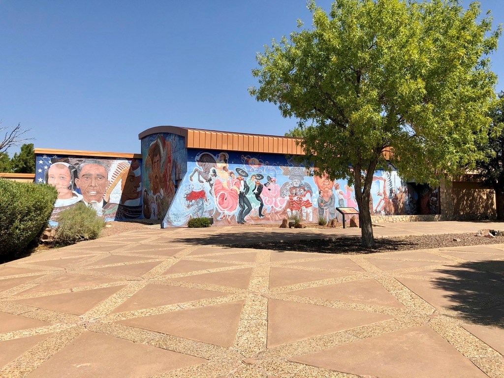 From across a paved plaza you can see a mural painted on three walls of a building. It shows vibrant, active figures depicting peoples and culture of the US-Mexico border.