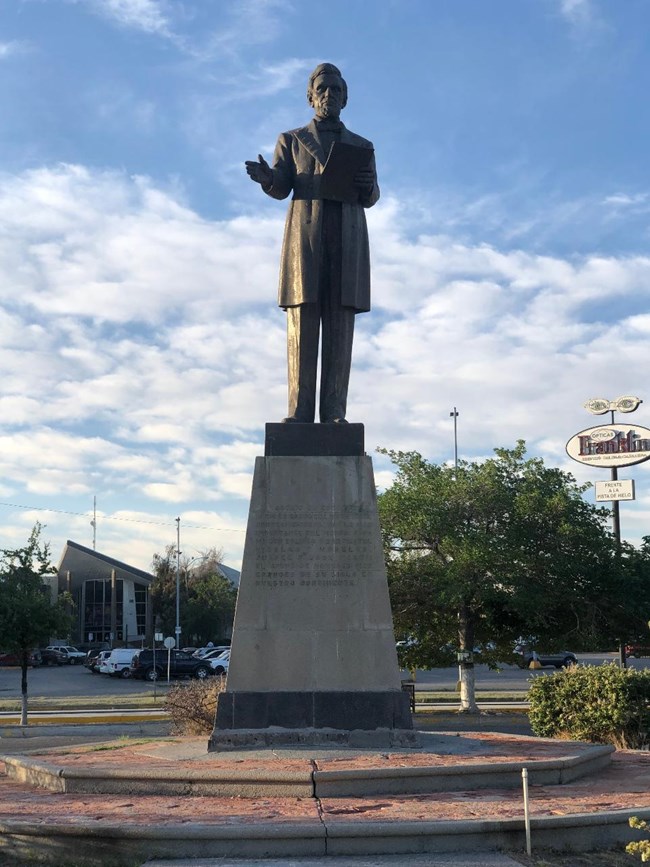 Bronze statue of President Lincoln on stone pedestal in road median. Lincoln his holding papers in left hand and gesturing with right.