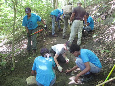 A group of young people wearing blue shirts work on a stretch of trail. Sunlight comes through thick forest overhead.