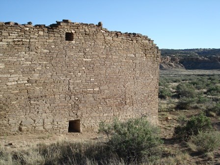 A stone and tan wall stands tall above the surrounding vegetation, with a glimpse of the desert background on the right side of the image.