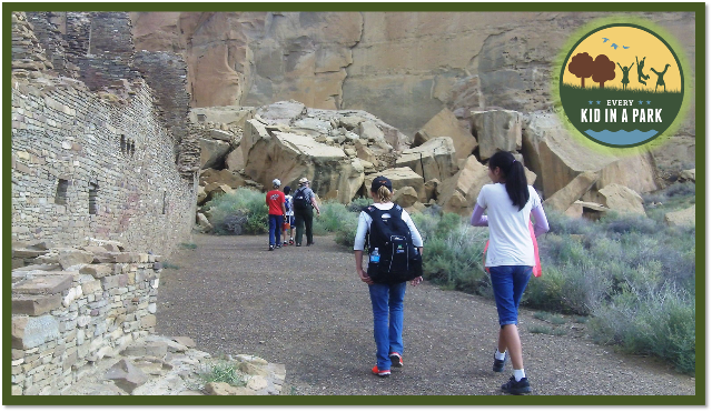Young children walk on a path away from the viewer with a park ranger in the distance, beside the tan remnants of ancient structures on the left.