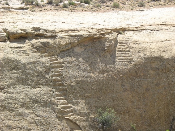 A tan mesa cliff face with carved steps and smaller handholds carved to either side of each step.