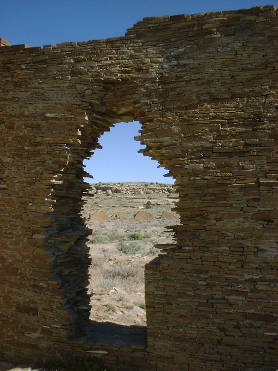 A tall masonry wall with a jagged doorway acts as a window to show the remains of masonry walls in the background, mixed among and blending into the desert landscape.