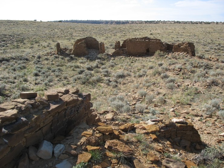 Remains of stone walls rise about the surrounding desert landscape, with vegetation reclaiming the area around the crumbling walls.