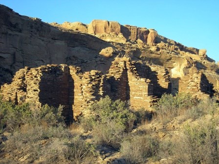 Remnants of tan stone-built walls rise about the green vegetation in front of the mesa wall.
