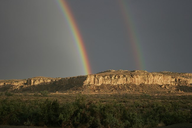 Dark clouds highlight a double rainbow rising high above the mesa and green vegetation of the canyon below.