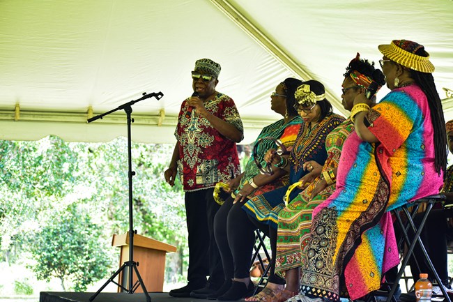 A man wearing sunglasses sings. He is wearing brightly colored clothes. Behind him, others are dressed similarly, seated and either singing or playing music.
