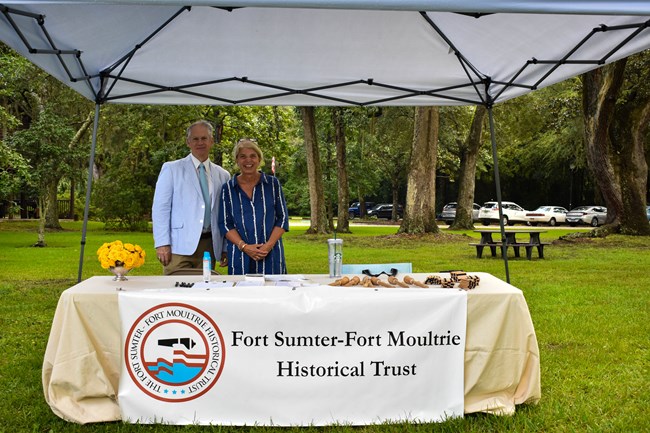 A man wearing a blue seersucker jacket and a woman wearing a blue dress stand under a white tent. The banner in front of them reads "Fort Sumter - Fort Moultrie Historical Trust"