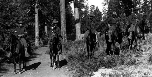 Black soldiers atop horses with rifles slung across their backs in a forested area