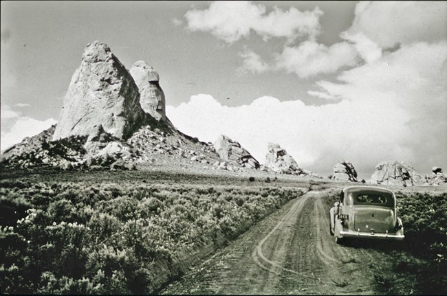 Black and white photo showing an old model car along a dirt road in front of towering granite spires.