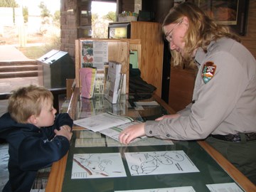 Ranger at information desk helping young person. Both are looking at a book on the desk.