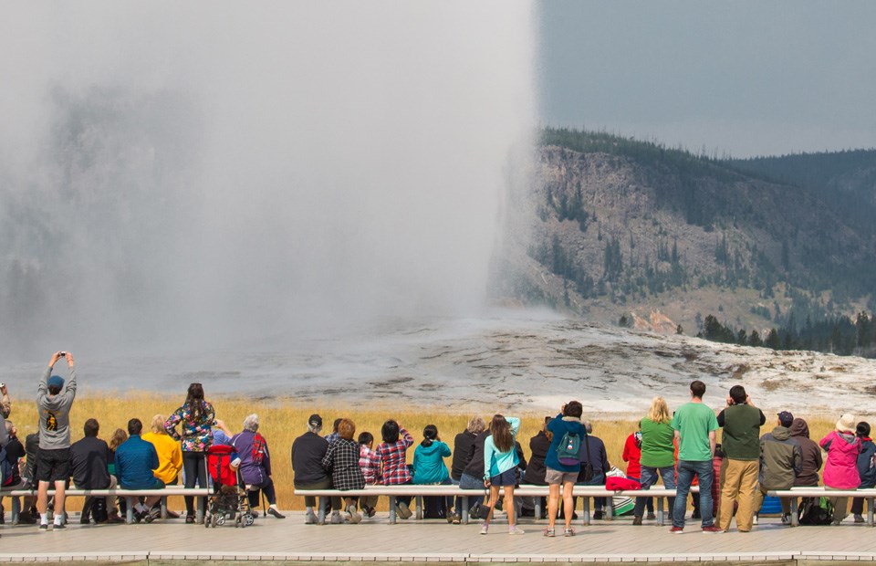 People stand on a boardwalk and watch steam and water gush into the air.
