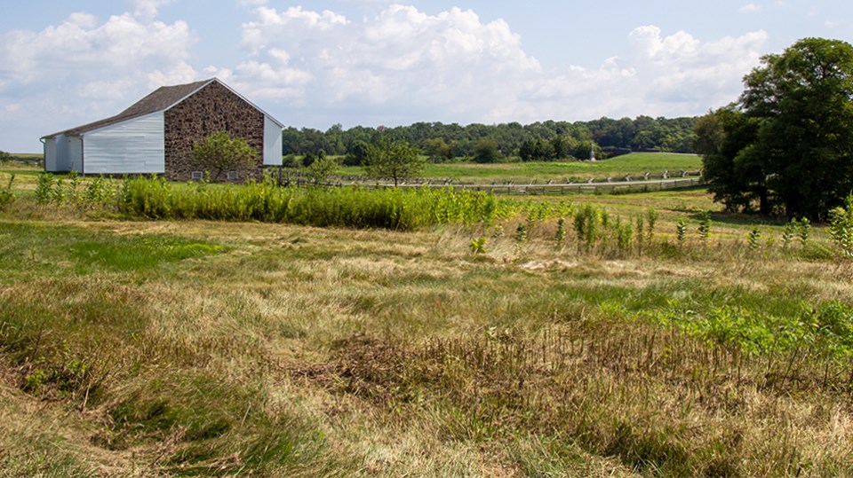 Two men look over a field at a large stone barn, a wagon shed, and a house.