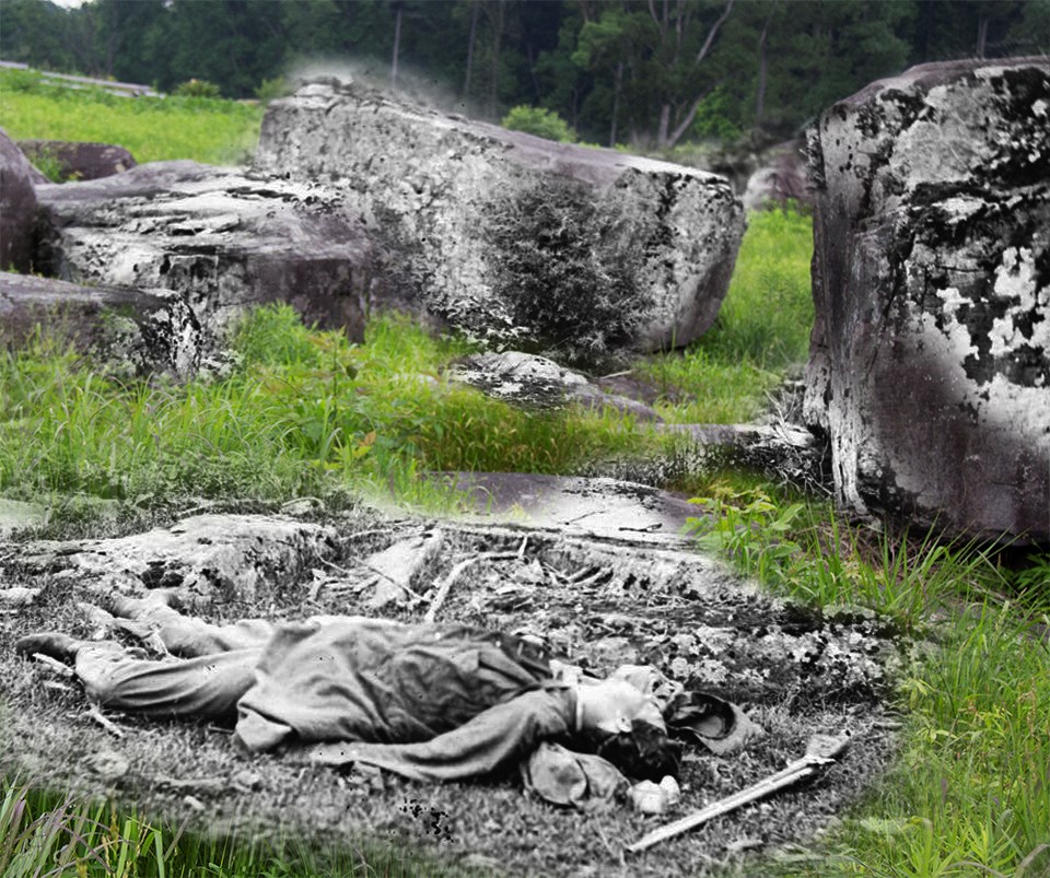 A dead Confederate soldier lies amidst debris near Devil’s Den, with a gun and hat near his head.