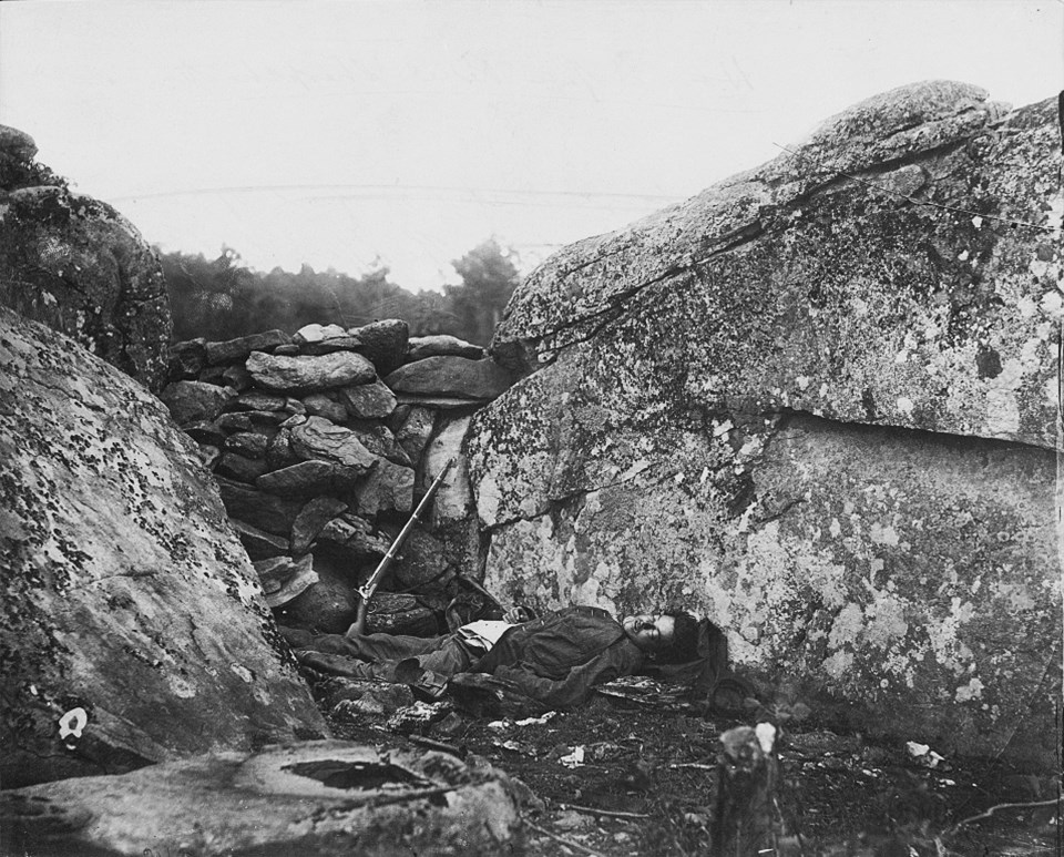 A dead Confederate soldier lies behind a stone fortification, a gun propped against the rocks next to him.