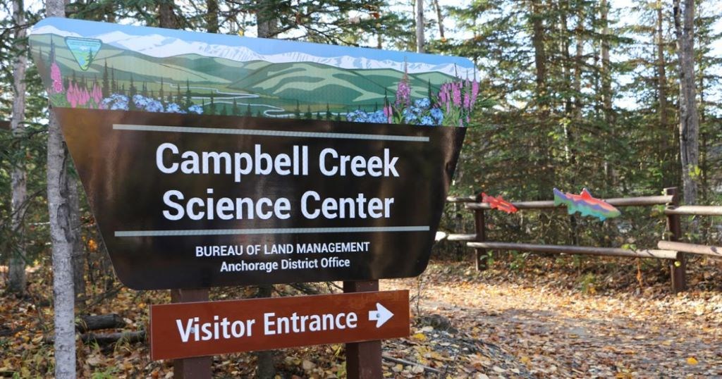 Close-up of sign that reads "Campbell Creek Science Center" with a smaller sign below reading "Visitor Entrance" and and arrow pointing to right; outdoor setting.