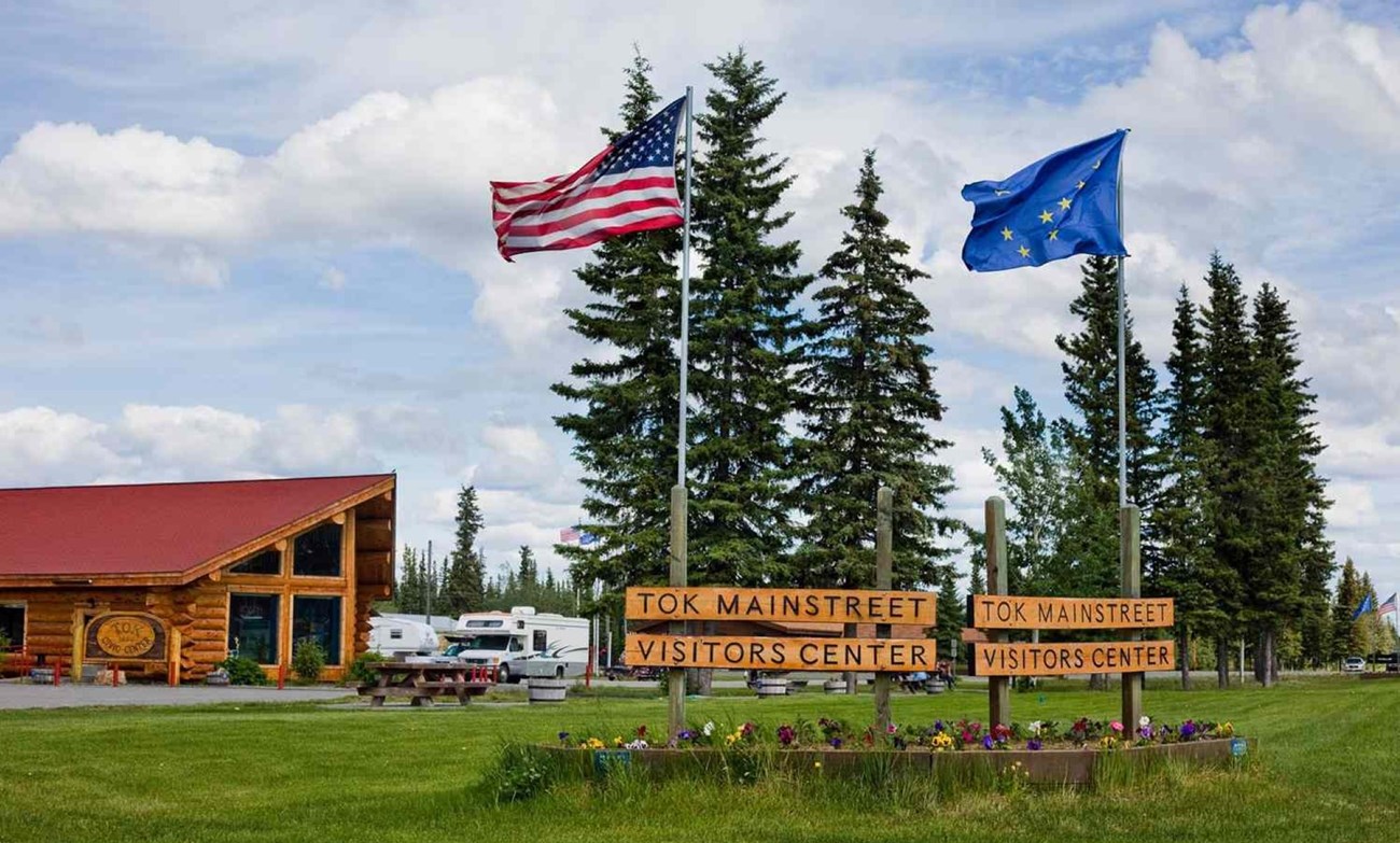 Outdoors; wooden building on left, 2 flags and 4 wooden signs in middle foreground, sky above and grass below.