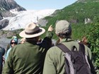 a ranger next to a visitor points towards a mountain slope with a glacier in the background.