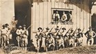 A row of men in hats and light shirts stand with rifles in front of a small clapboard shack.
