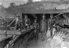 Young men in miners gear stand in front of mine carts and the opening to a mine shaft. 