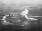 Aerial shot bombing of ships in lagoon, large plumes of smoke rising, landforms in background.
