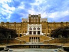 Front of a high school made of brown brick that rises to a high point in the middle with stairways.