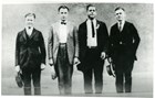 A row of men in suits and ties with hats in their hands stand on a street.