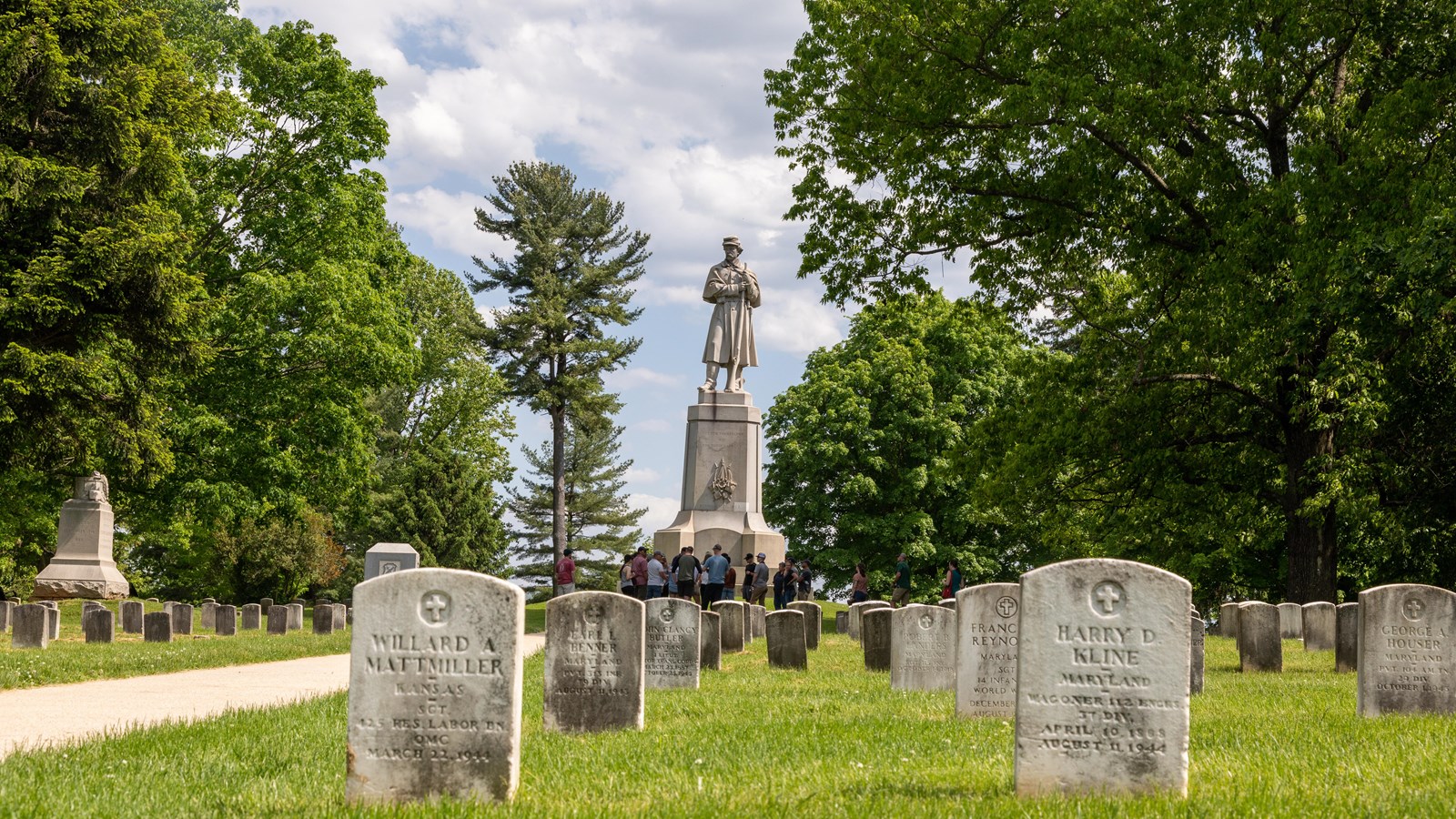 A monument of a private stands among graves in a cemetery. Students stand in a group in front of it.