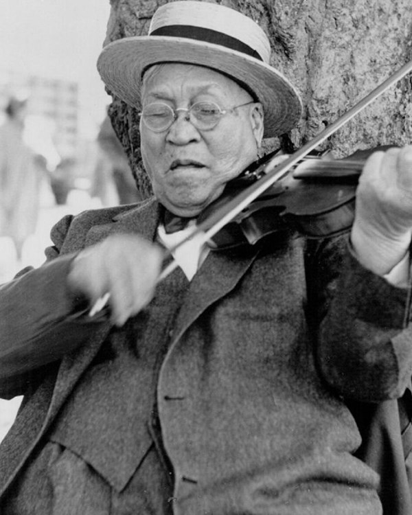 Black and white image of a man seated fiddling vigorously. He is wearing a three piece suit and hat