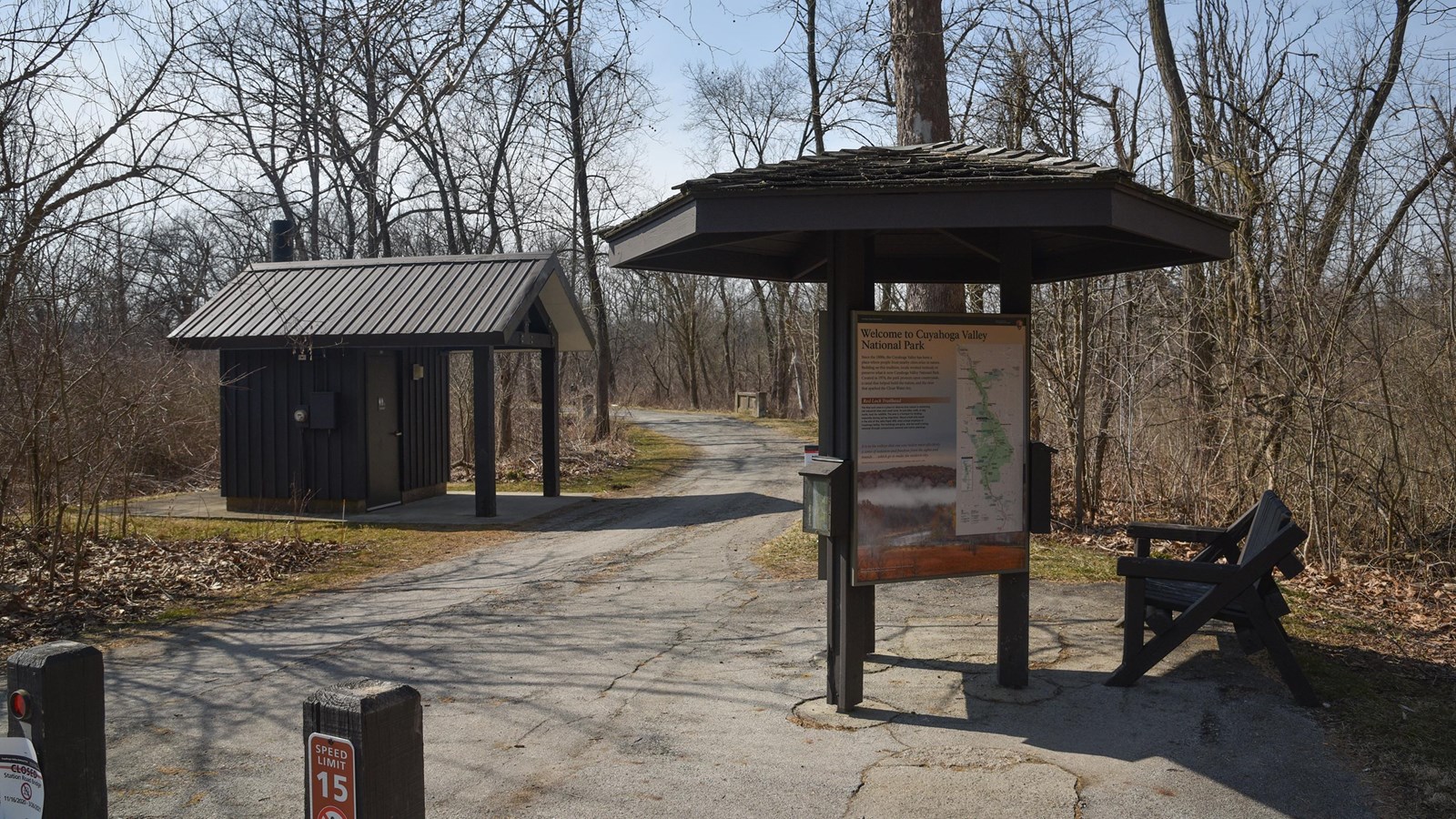 Paved trail passes a 3-sided kiosk and brown bench at right and a brown restroom structure at left.
