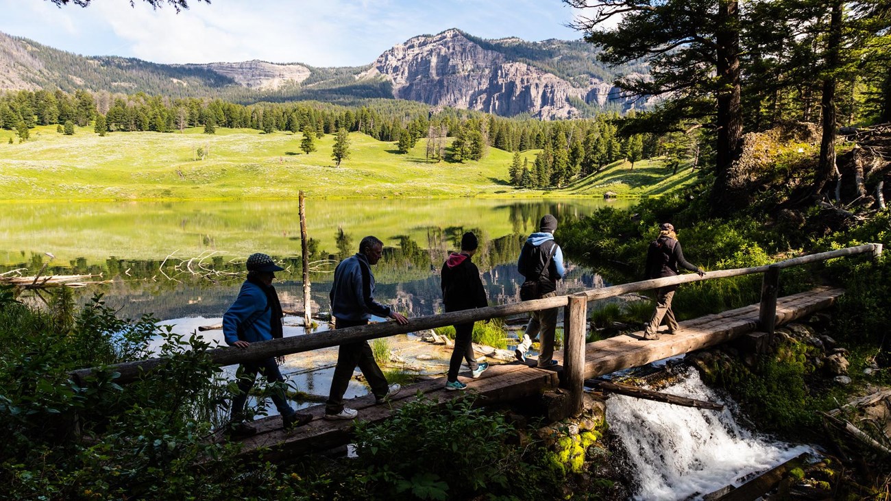 Hikers walk across a bridge at the outlet of a small lake surrounded by rolling hills.