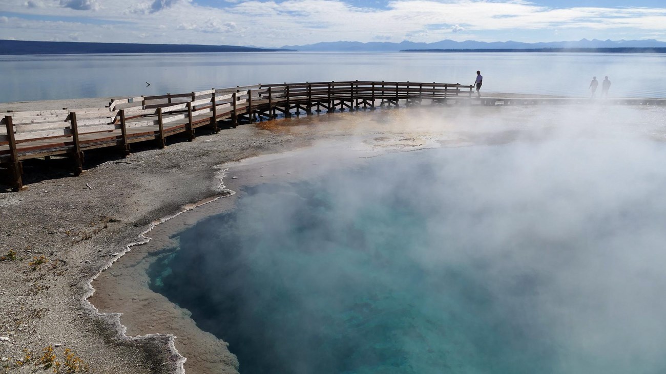 A turquoise hot spring pool surrounded by a boardwalk sits above a large lake.