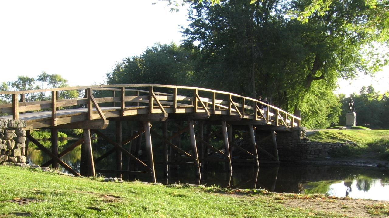 Arched wooden bridge with field stone abutments spans a slow river bounded by green embankments