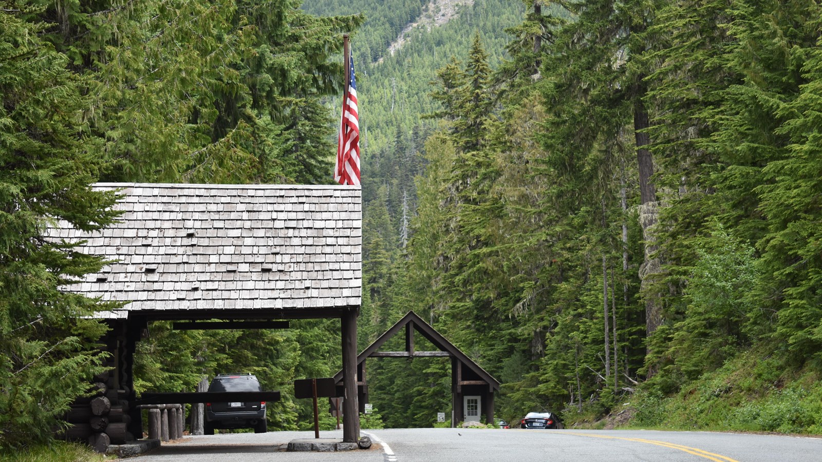 An American flag over a rustic porch roof with the high peaked roof of the entrance station beyond.